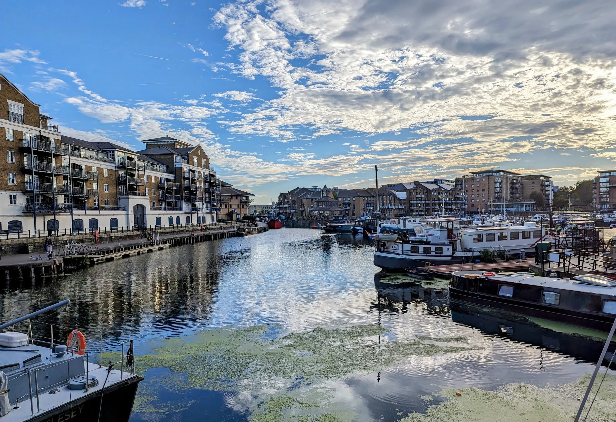 Limehouse Basin on a bright afternoon, narrowboats and cruisers moored along the pontoons and brick warehouse flats around the water