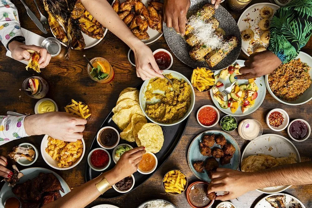 A wooden table shot from above covered in Caribbean dishes - grilled corn, wings, a curry, fried plantain, macaroni pie, fritters and small bowls of sauces - with a dozen hands reaching across it