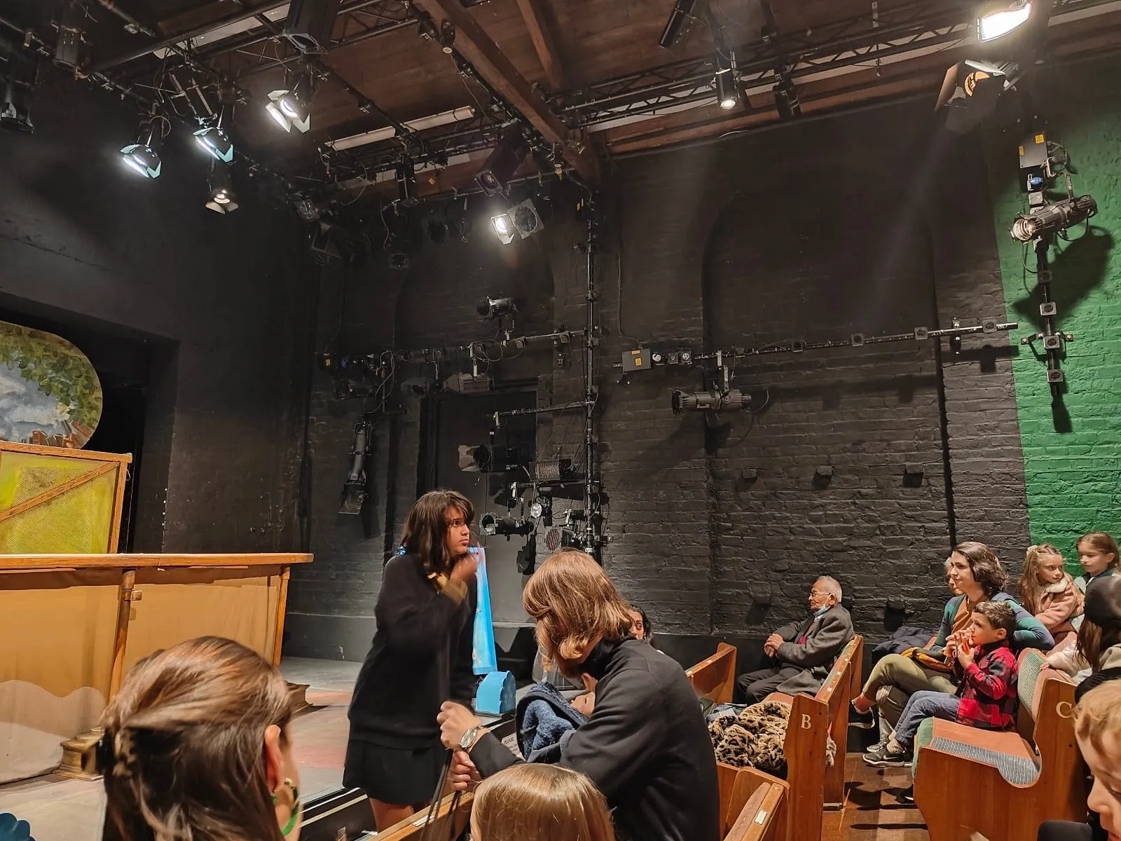 Children and adults on wooden pews at Little Angel Theatre, a few feet from a small stage with a painted puppet booth, under a rig of stage lights