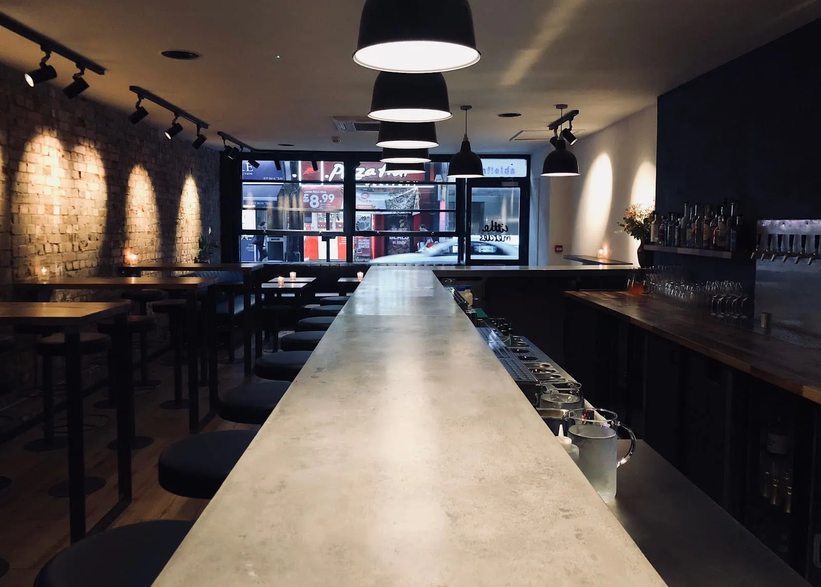 A long concrete bar counter with stools along one side, black pendant lamps overhead, high tables with candles against a brick wall, and the street through the front window