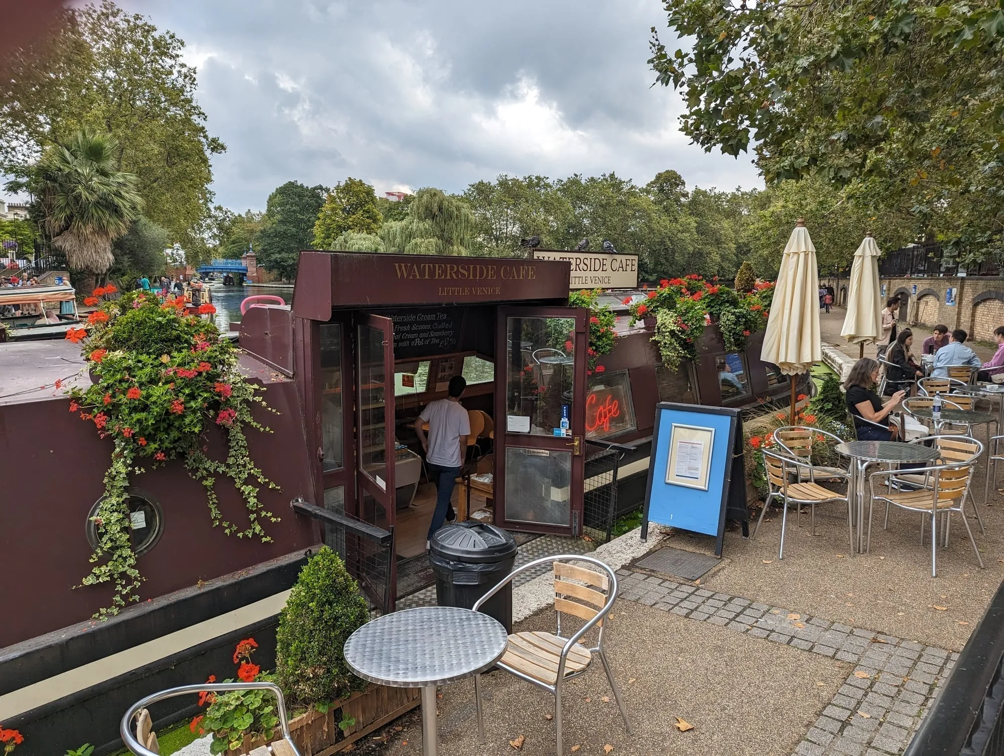 The Waterside Cafe at Little Venice, a maroon narrowboat with red geraniums serving customers at canalside tables