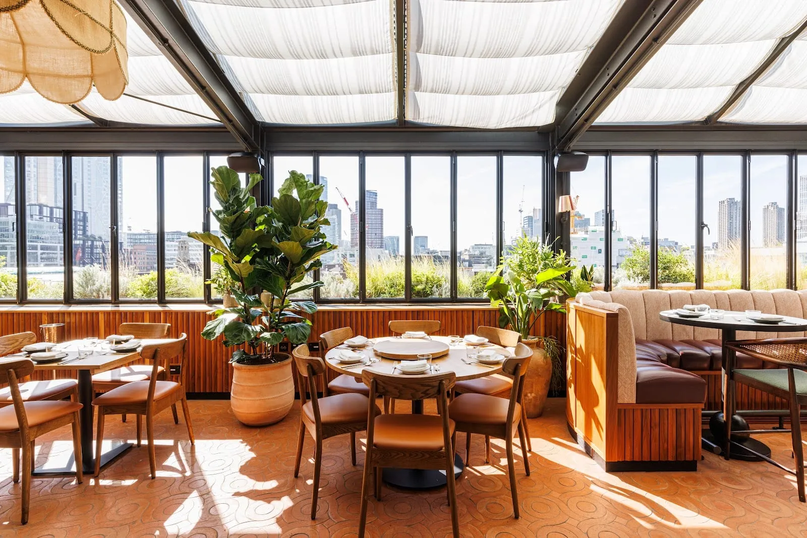 A glass-roofed rooftop dining room with white fabric shades, terracotta floor tiles, large potted fig trees and an east London skyline through the windows