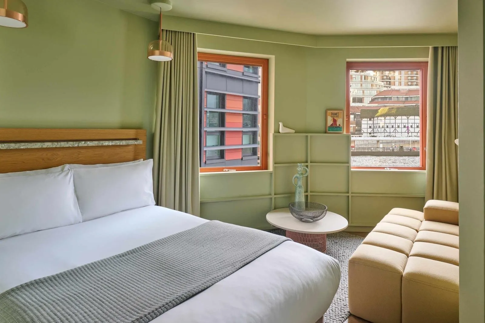 A pale green room at Locke at Broken Wharf with a double bed, an oak headboard, a padded sand-coloured sofa and two orange-framed windows, one looking across the Thames to Shakespeare's Globe