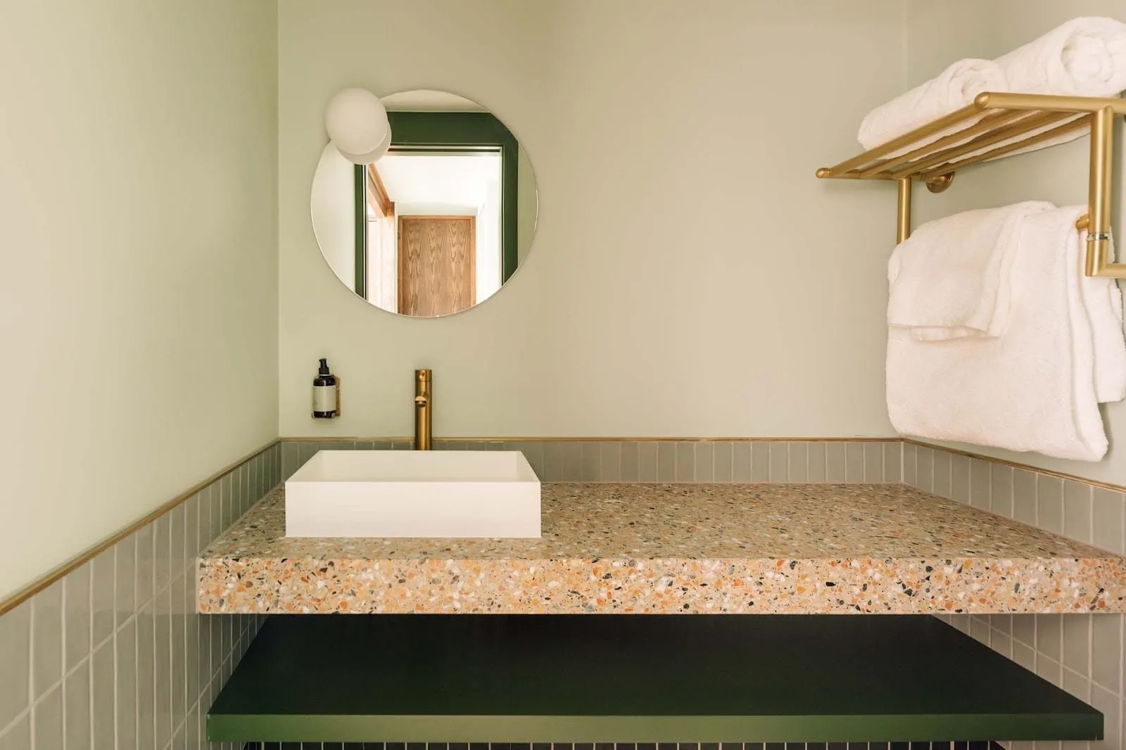A basin at Locke London Canary Wharf: a white square sink on a pink terrazzo counter, sage tiles with a brass trim, a round mirror and a brass towel rail