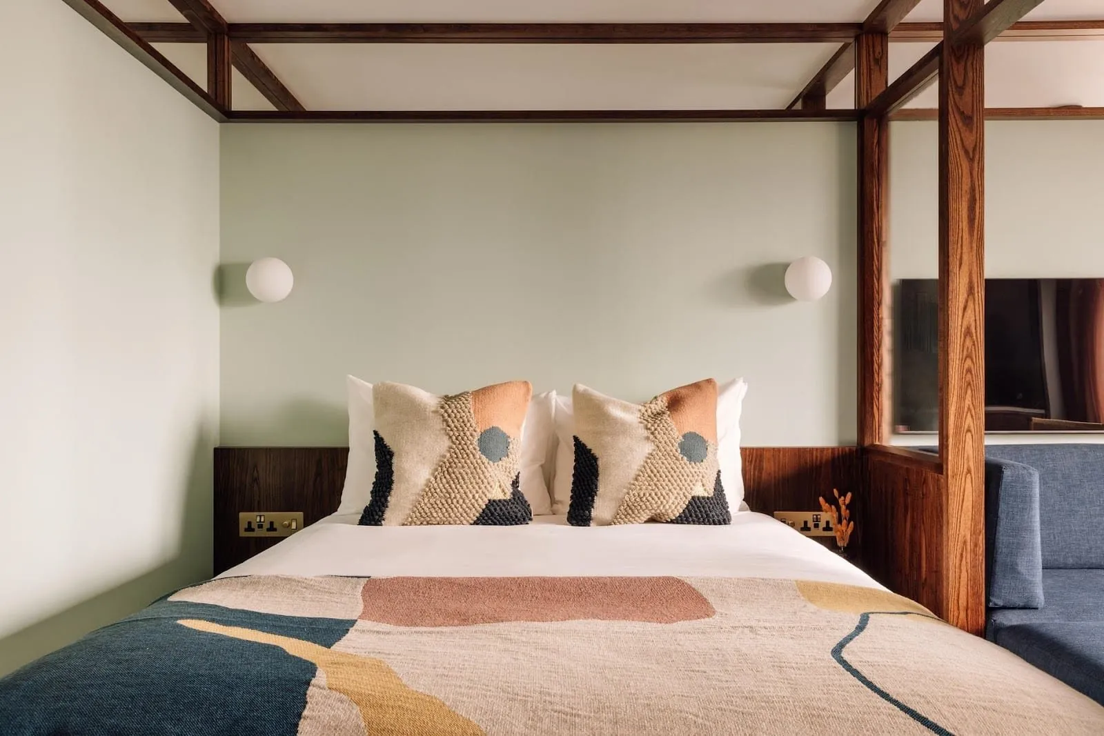 A studio at Locke London Canary Wharf: a double bed inside a dark timber four-poster frame, sage-green walls, globe wall lights, a woven throw and a blue sofa beside the bed