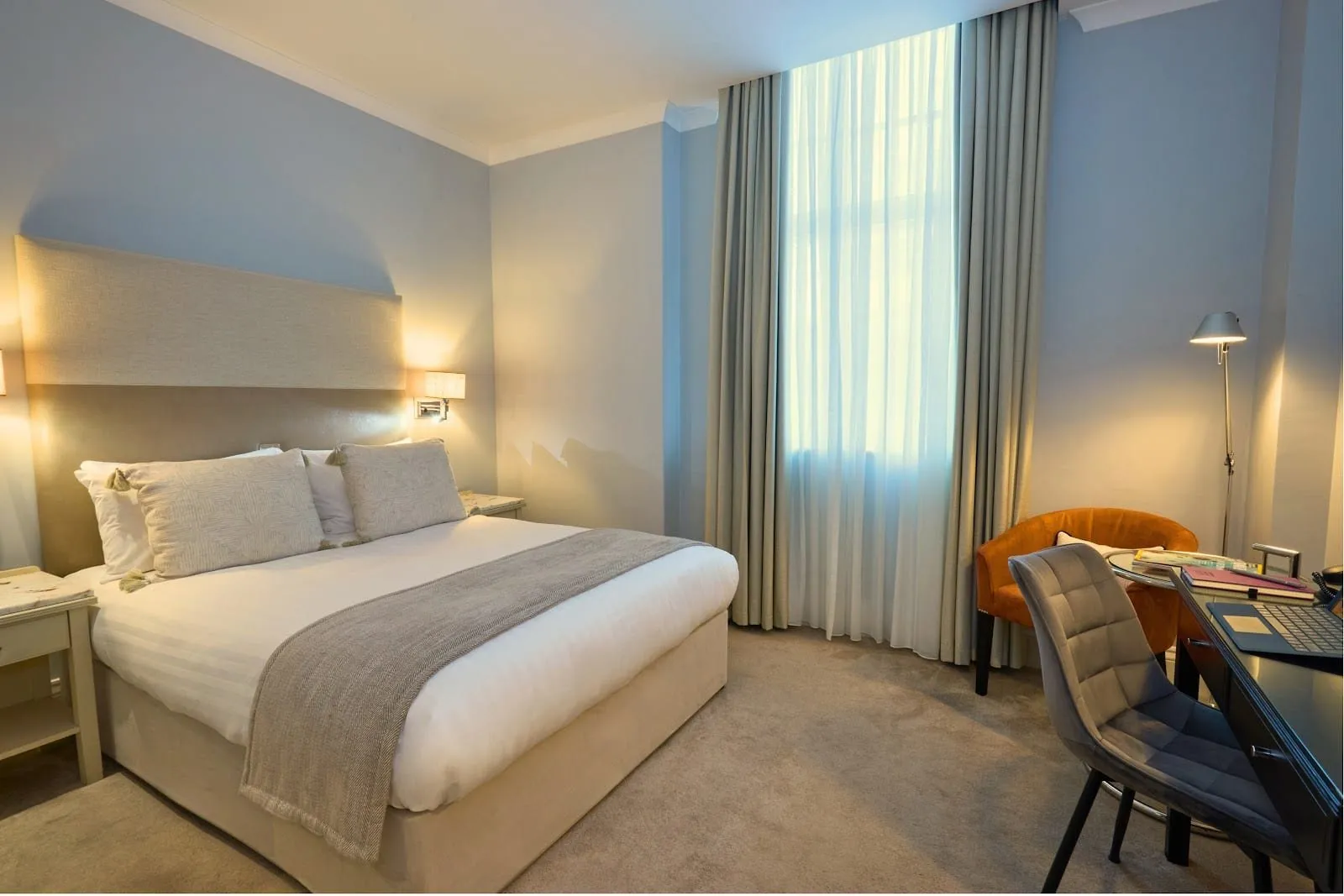A double room at London Bridge Hotel with a tall upholstered headboard, pale blue walls, a tall window with full-length curtains, an orange tub chair and a desk