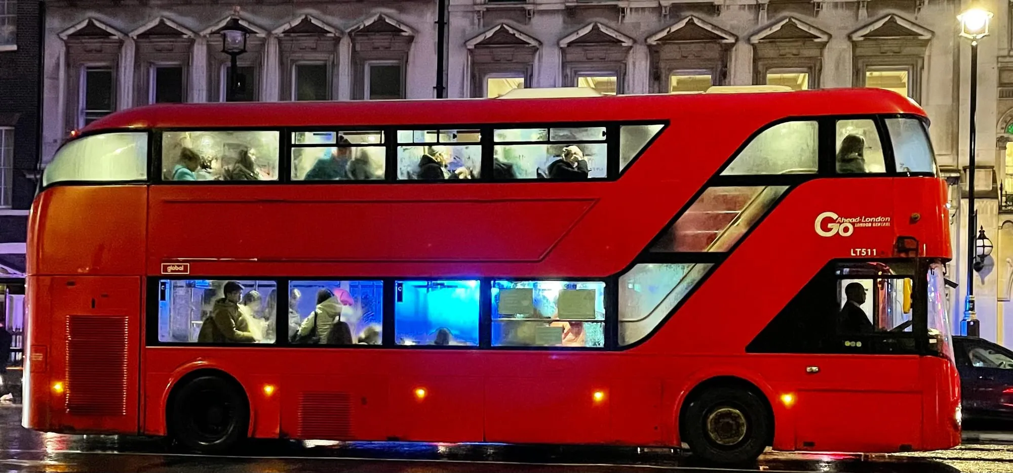 A brightly lit red London double-decker bus with passengers aboard, driving through the city on a wet street at night