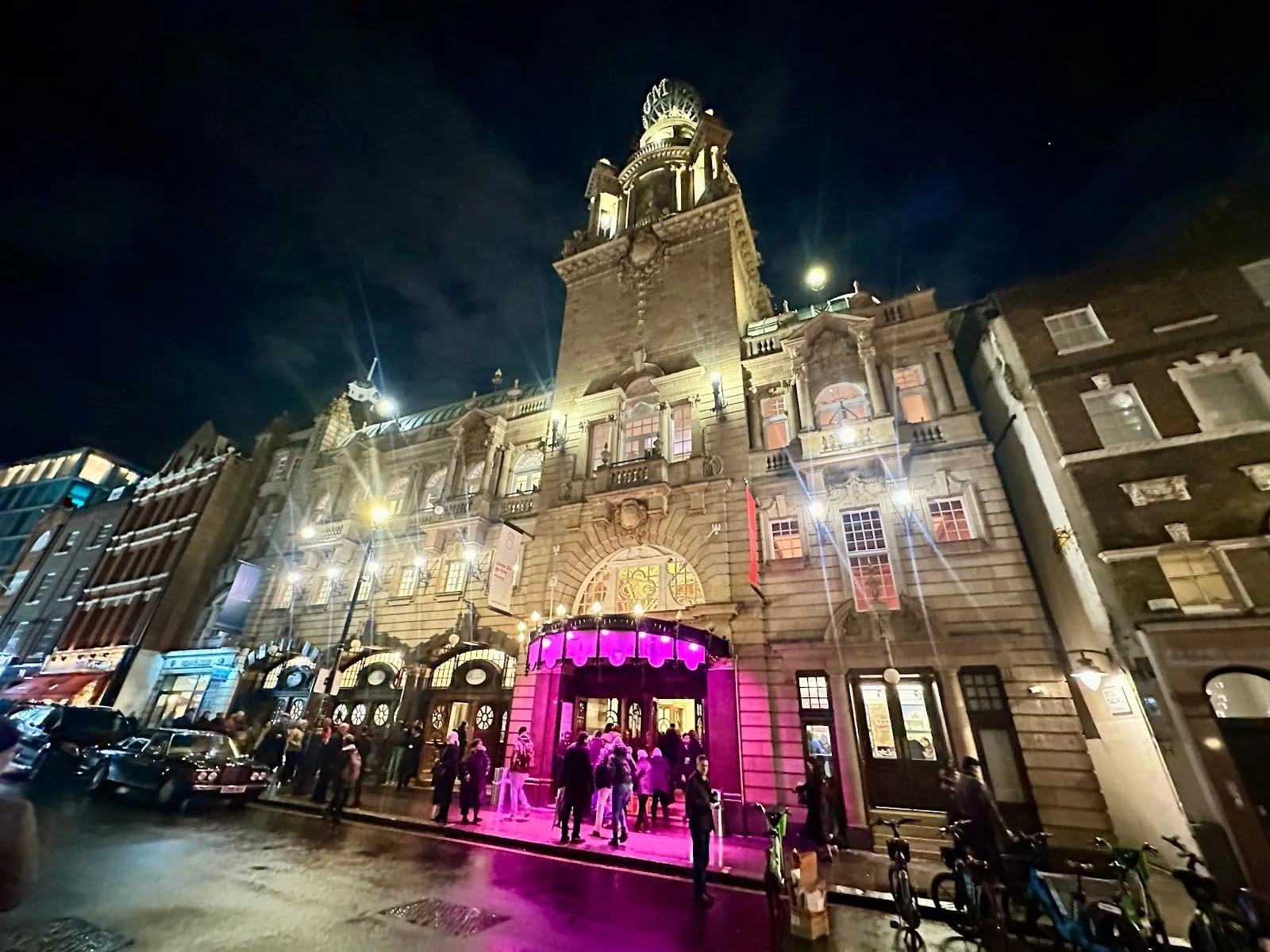 The London Coliseum at night, its tower topped by a lit globe, with an audience going in under a pink-lit entrance canopy