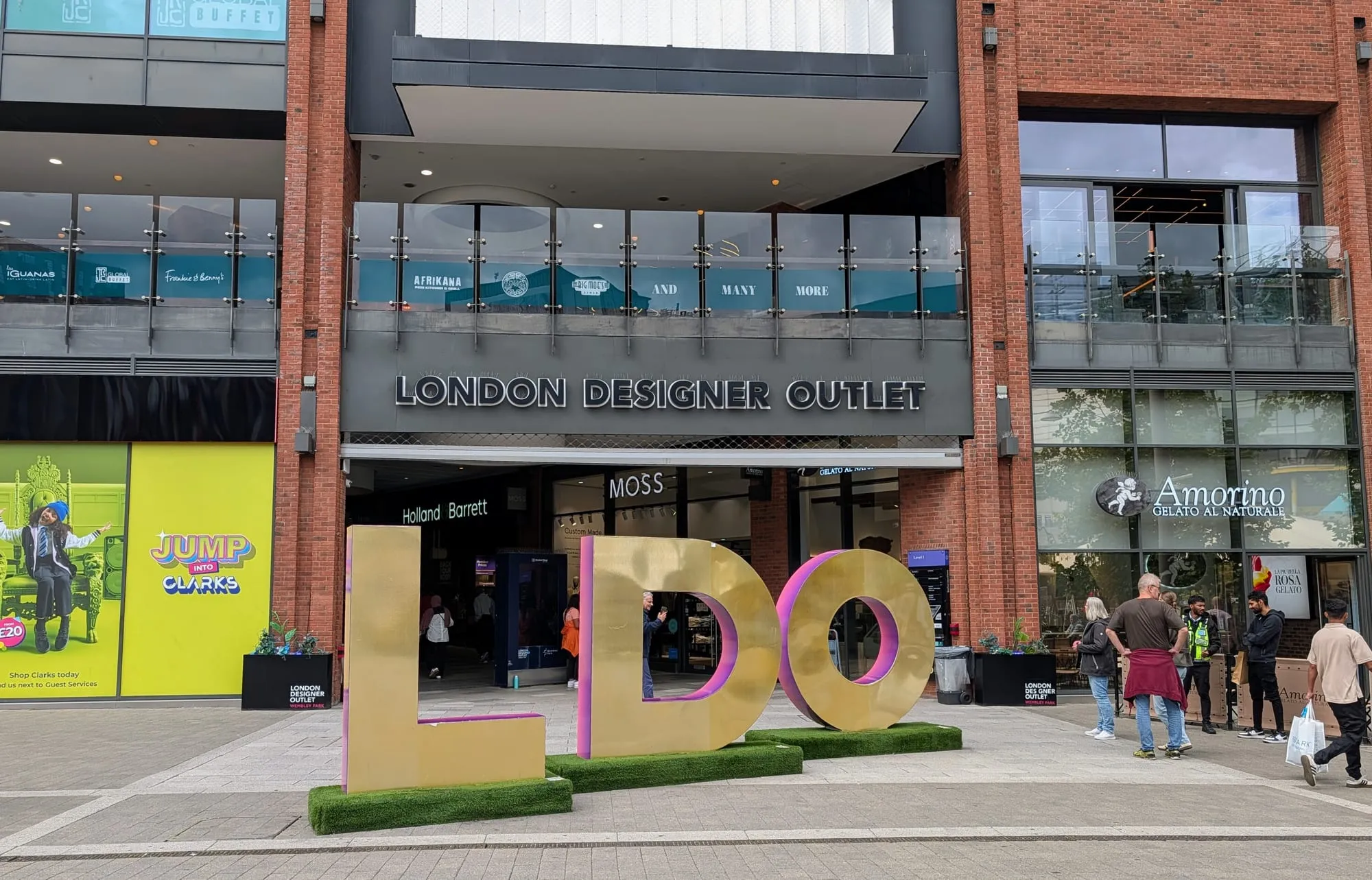 The entrance to the London Designer Outlet at Wembley Park, with giant gold LDO letters standing on grass plinths in front of a red brick and glass frontage, Holland & Barrett and MOSS inside the doorway and an Amorino gelato shop to the right