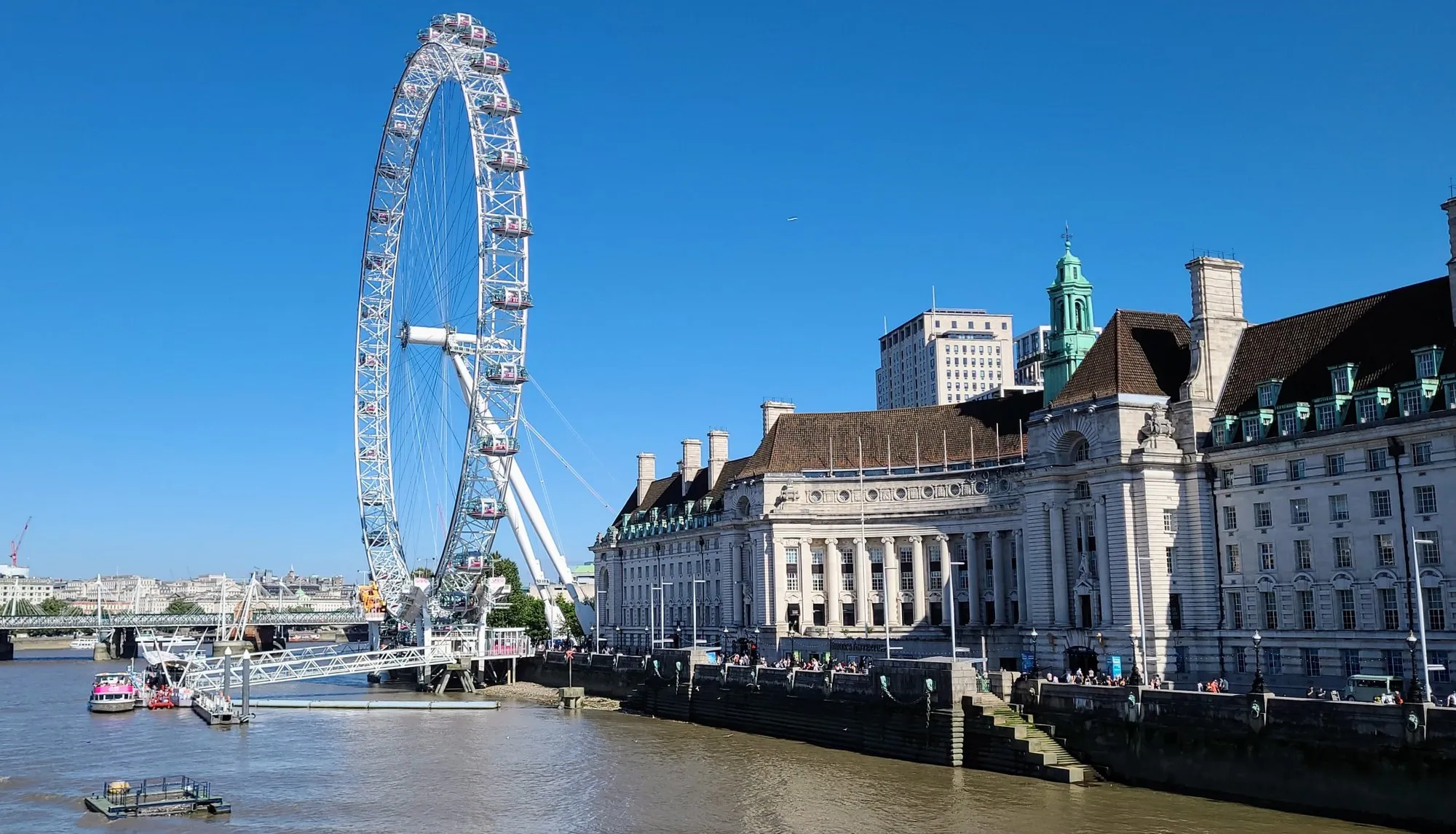 The London Eye and County Hall across the Thames on a clear summer day, with the Golden Jubilee Bridges and a river boat at the pier