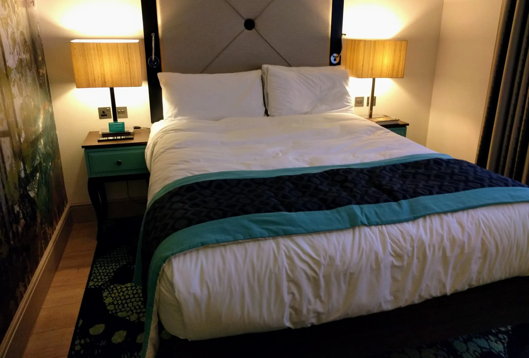 A London hotel room with a double bed, a patterned throw, teal bedside tables and lamps lit either side of an upholstered headboard