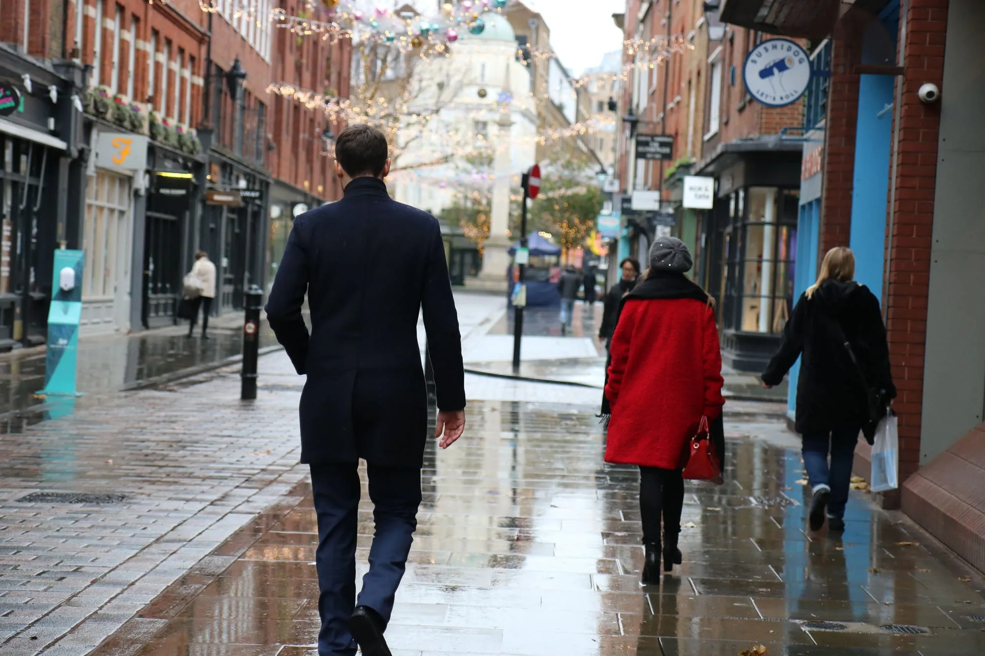 People walking through Seven Dials in London on a wet day, festoon lights overhead and the pavement reflecting