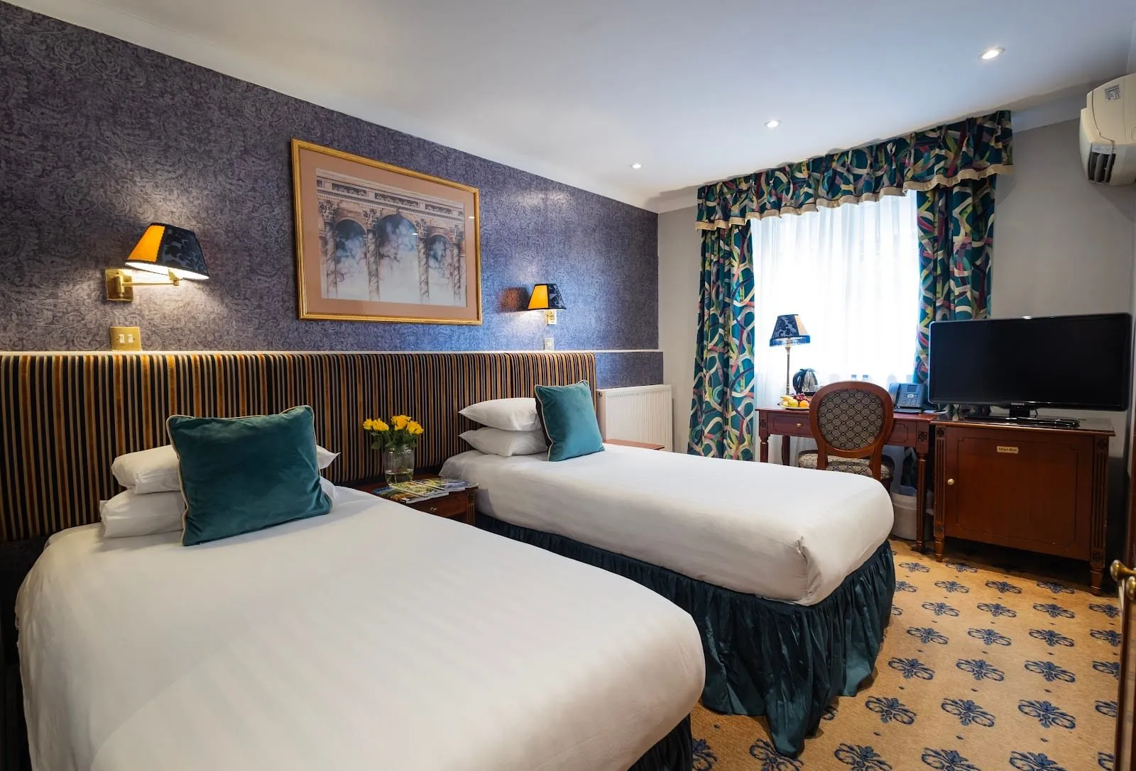 A twin room at London Lodge Hotel with two single beds against a striped velvet headboard, teal cushions, patterned curtains, a mahogany desk and a wall-mounted air-conditioning unit