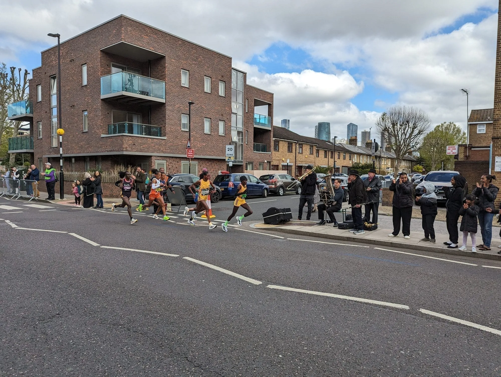 The elite women's field running past spectators and a brass band on a London street during the TCS London Marathon