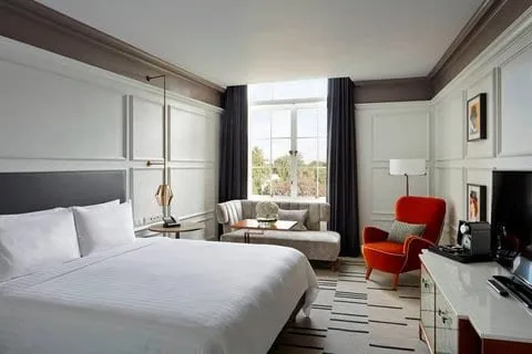 A room at the London Marriott County Hall with white panelled walls, a grey headboard, an orange armchair, a grey sofa and a tall window onto trees