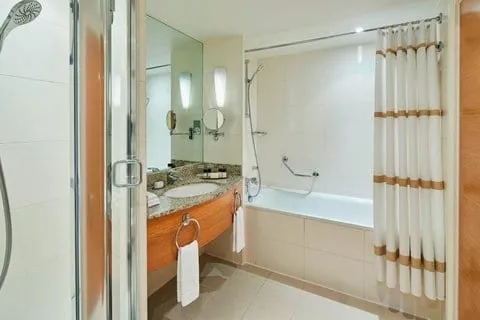 A bathroom at the London Marriott Hotel Canary Wharf with a bath behind a striped curtain, a separate glass shower cubicle, and a granite vanity over a wood cabinet