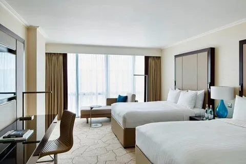 A room at the London Marriott Hotel Canary Wharf with two double beds against a tall upholstered headboard, a glass desk, and a chaise under floor-to-ceiling windows with sheer curtains
