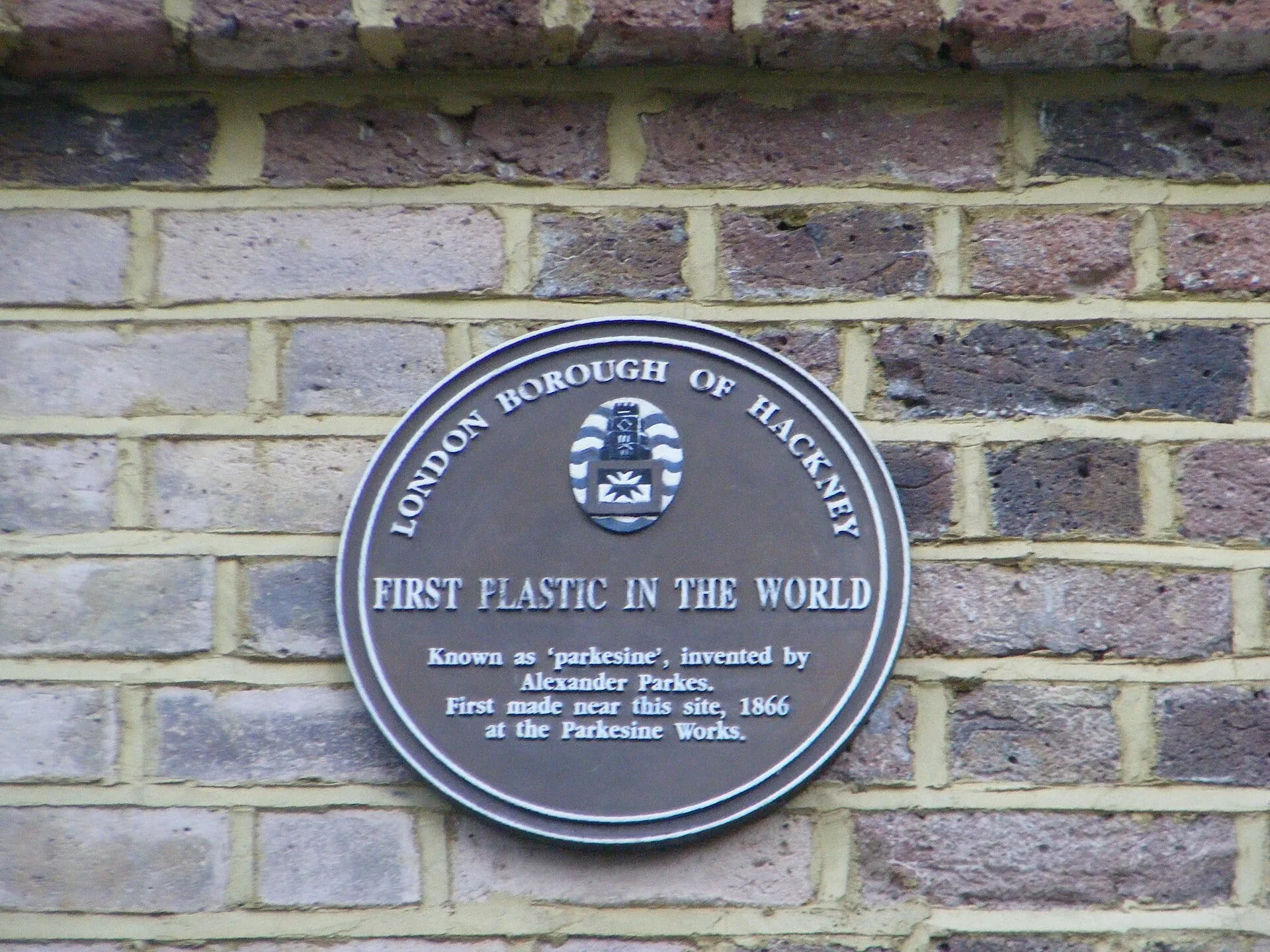 A brown London Borough of Hackney plaque on a brick wall, reading FIRST PLASTIC IN THE WORLD