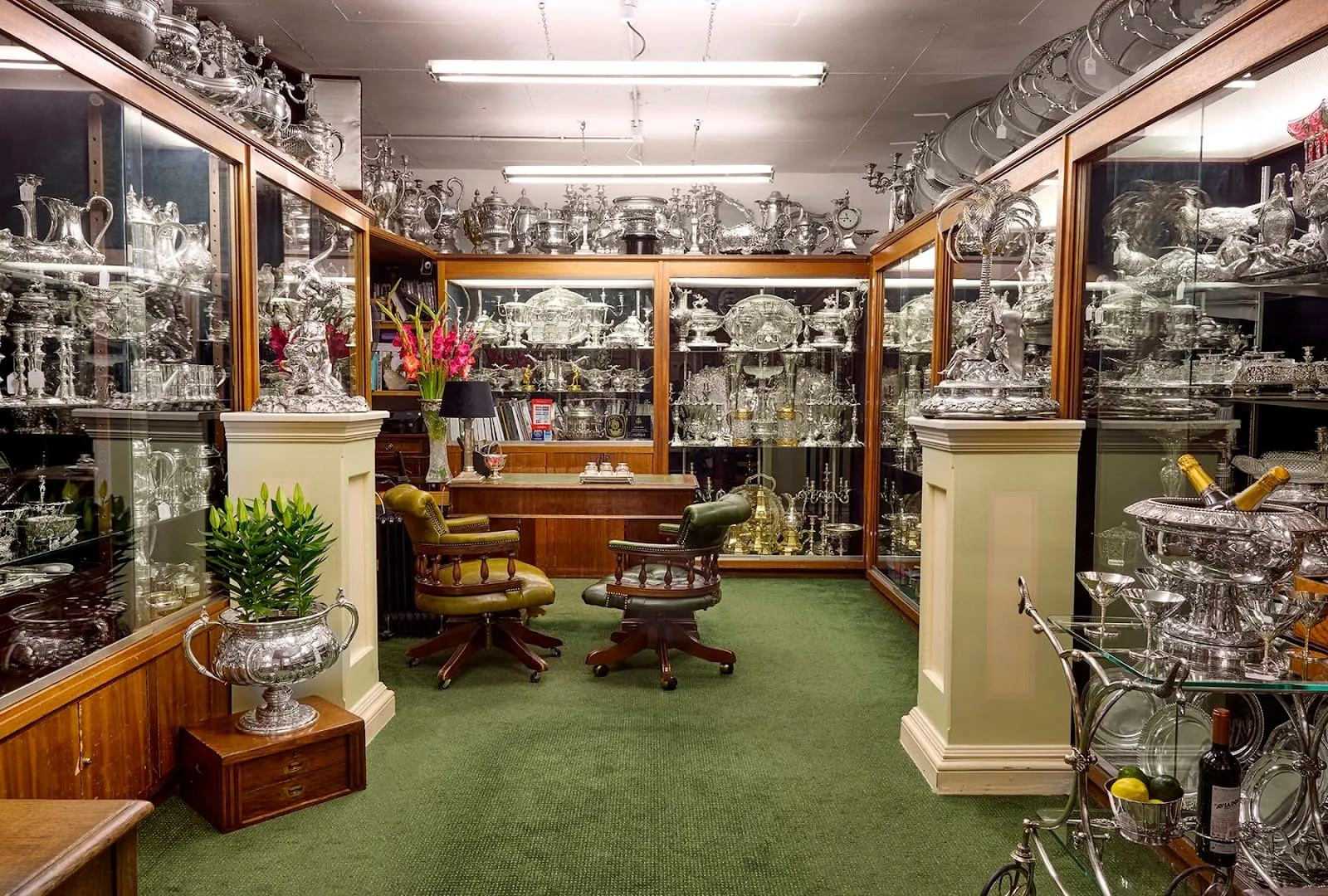 A dealer's shop in the London Silver Vaults, with glass cabinets and the tops of the walls packed with silver teapots, trophies and candelabra above a green carpet