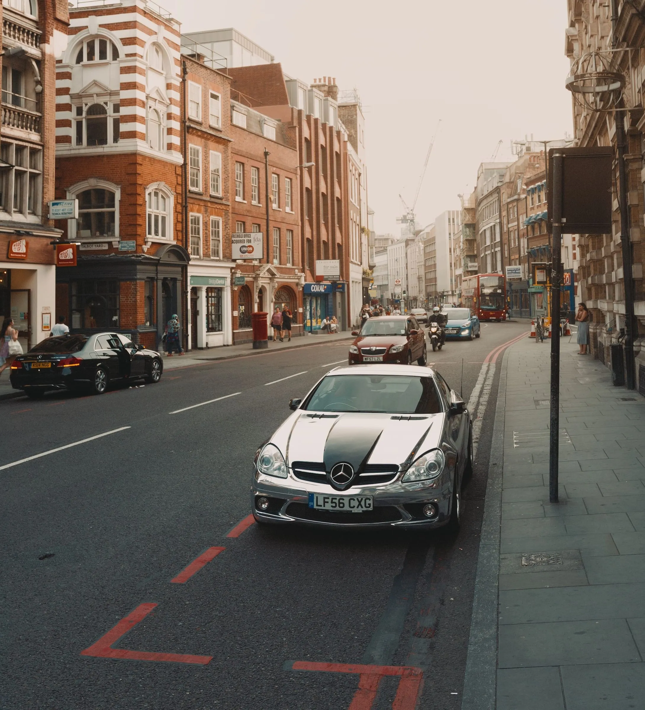 A London street of red-brick terraces with cars parked along a red route and traffic heading towards the city