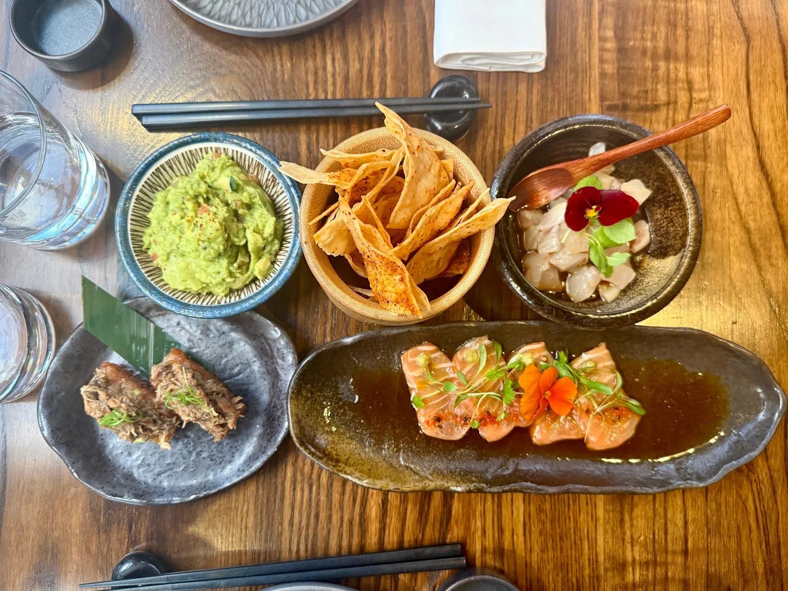 A table at Los Mochis seen from above: guacamole, tortilla chips, a white fish ceviche with an edible flower, salmon tiradito in a dark dressing and pulled duck on crispy rice, with chopsticks
