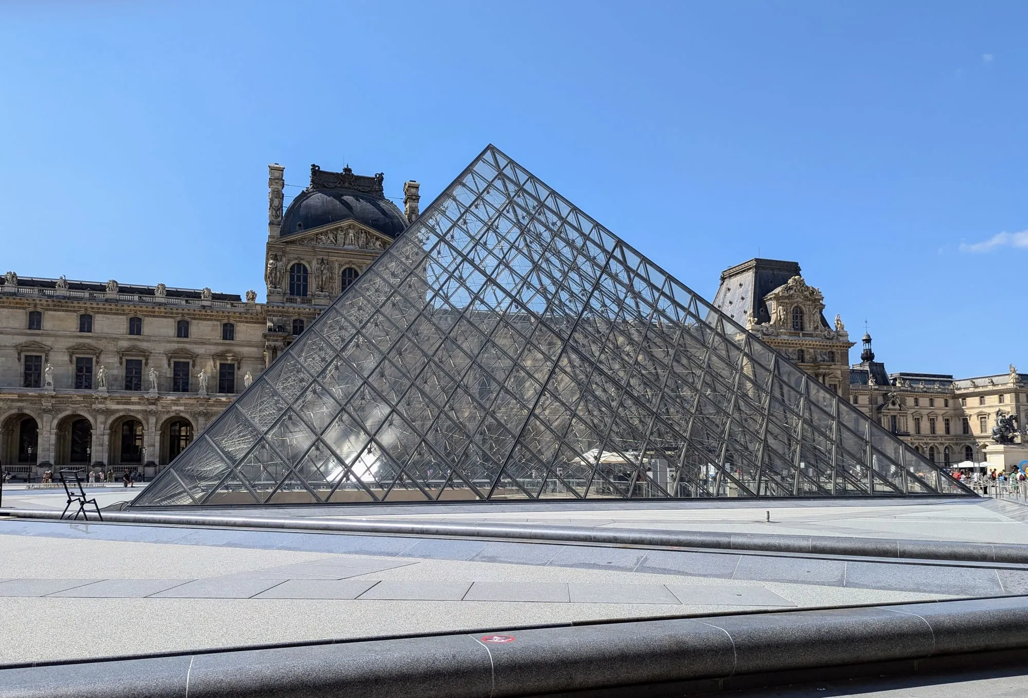 The Louvre's glass pyramid in its main courtyard, with the palace's carved stone wings behind it under a clear sky