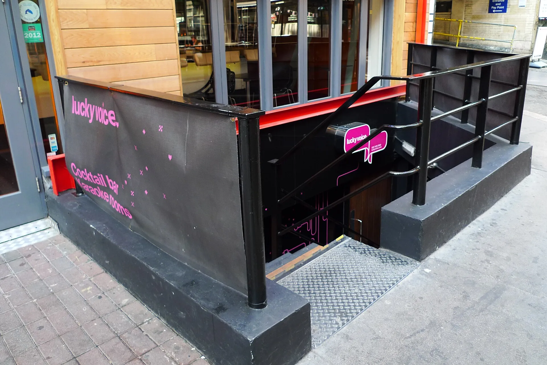 The basement entrance to Lucky Voice on Poland Street, with a pink Lucky Voice sign over the stairs and a banner reading cocktail bar, karaoke rooms