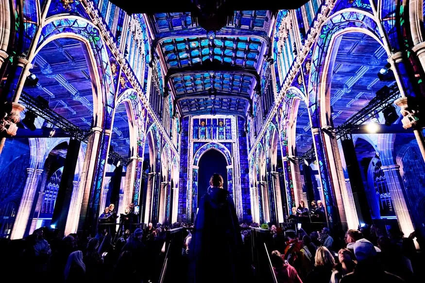 A cathedral nave washed deep blue by projected stained-glass patterns across every arch and the ceiling, with an audience seated below