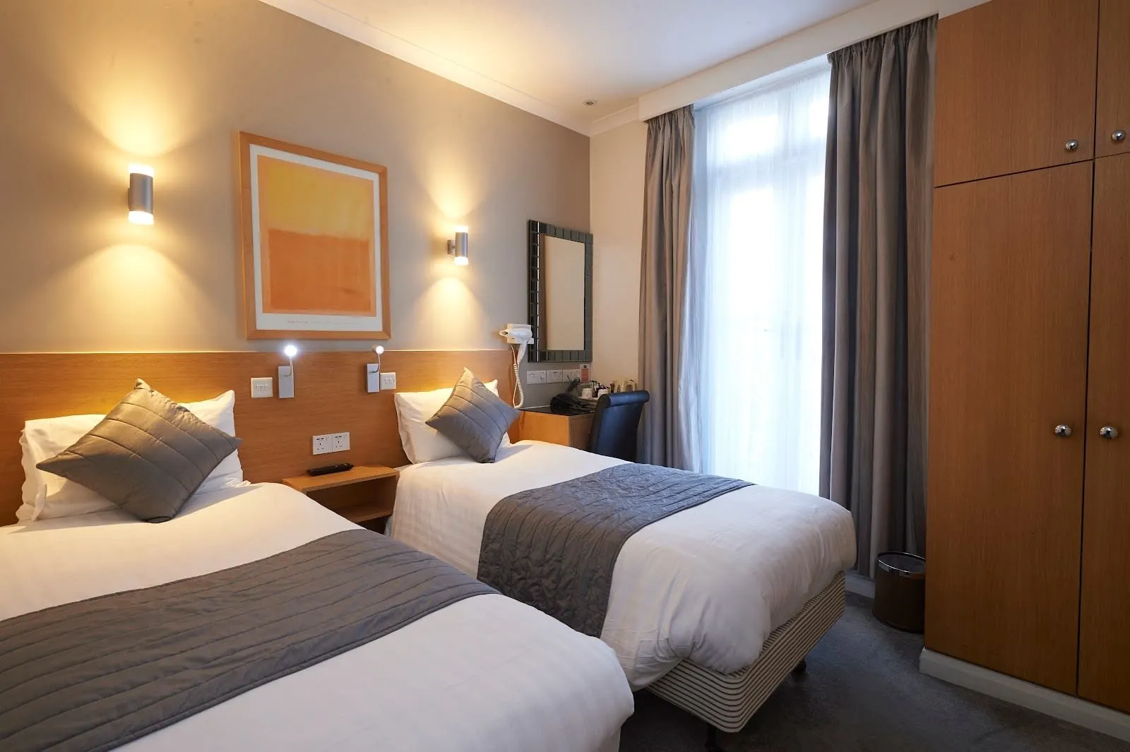 A twin room at Luna Simone with two single beds against a light wood headboard panel, grey bed runners, a tall window with grey curtains and a built-in wardrobe