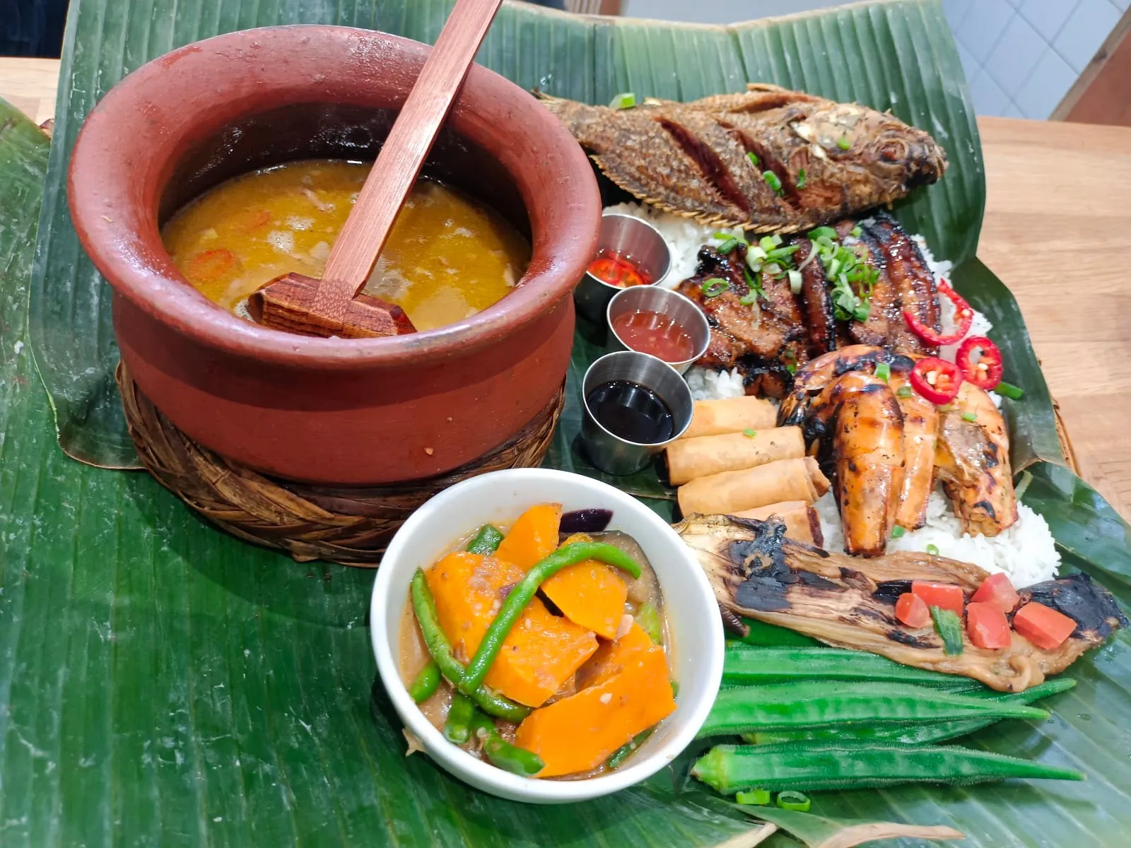 A banana leaf laid over a table, holding a clay pot of golden broth, a bowl of squash and green beans in a thick sauce, a whole fried fish and grilled pork belly on mounds of rice, grilled prawns, spring rolls, three dipping sauces, a charred aubergine and raw okra