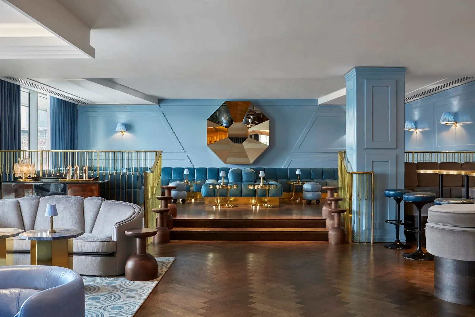 A raised lounge with blue velvet banquette seating around low brass-legged tables, grey armchairs in the foreground, a faceted brass octagonal mirror on a panelled blue wall, and a dark herringbone wood floor