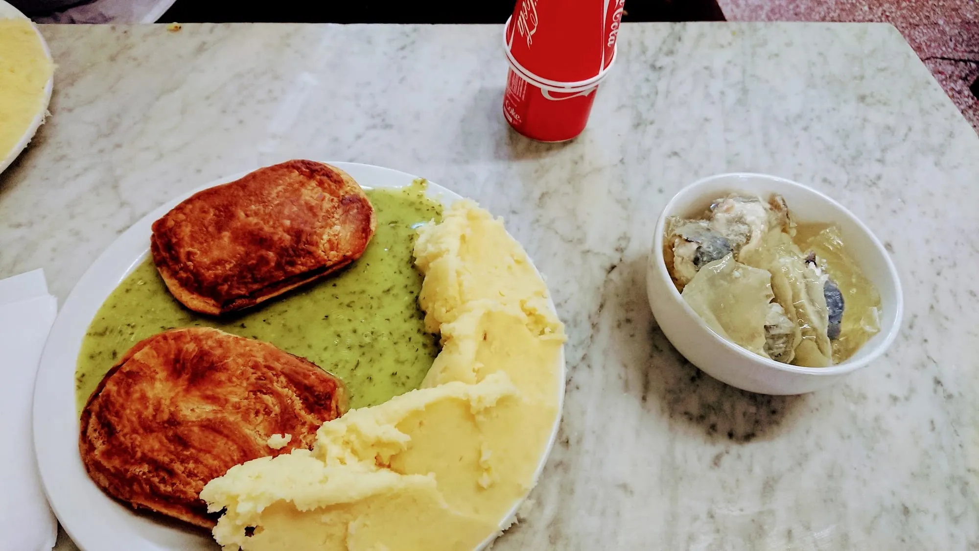 Two pies covered in green parsley liquor next to mashed potato and a bowl of jellied eels, on a marble table.