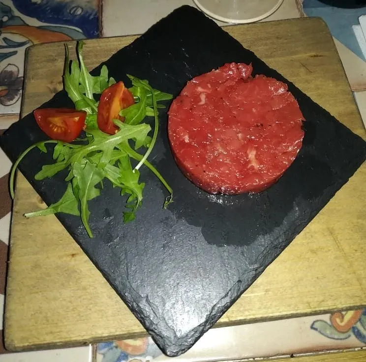 A disc of raw chopped Fassona beef on a black slate board, with rocket and two tomato wedges beside it