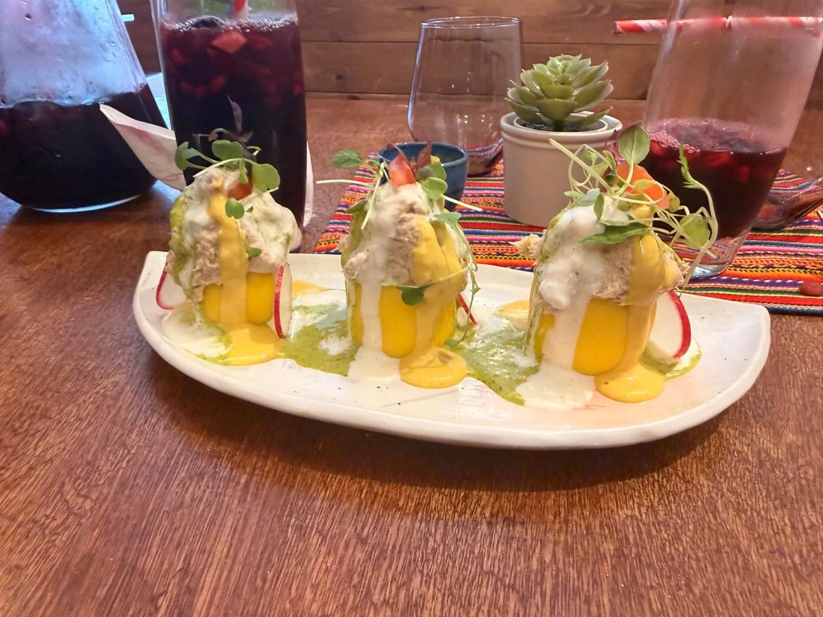 Three cylinders of bright yellow potato topped with a pale mayonnaise-dressed filling and micro herbs, on a white plate under yellow and green sauces, with jugs of dark purple chicha morada behind