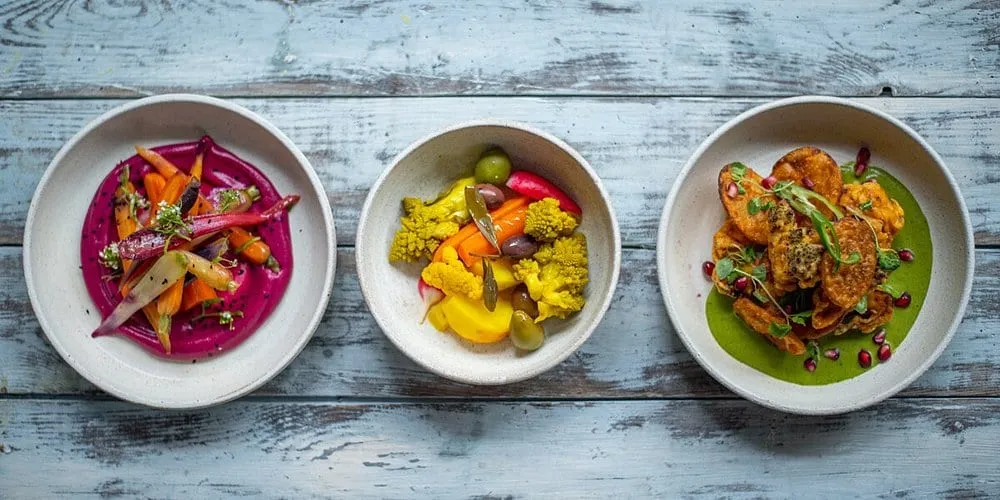Three bowls on weathered blue-grey wood: heritage carrots on a beetroot purée, pickled vegetables with olives, and fritters on a green herb sauce scattered with pomegranate