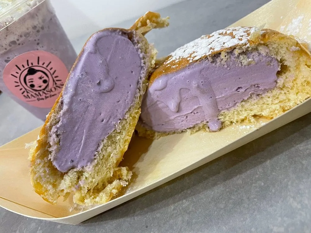 A bilog cut in half on a wooden tray — a toasted milk bun filled with bright purple ube ice cream and dusted with icing sugar