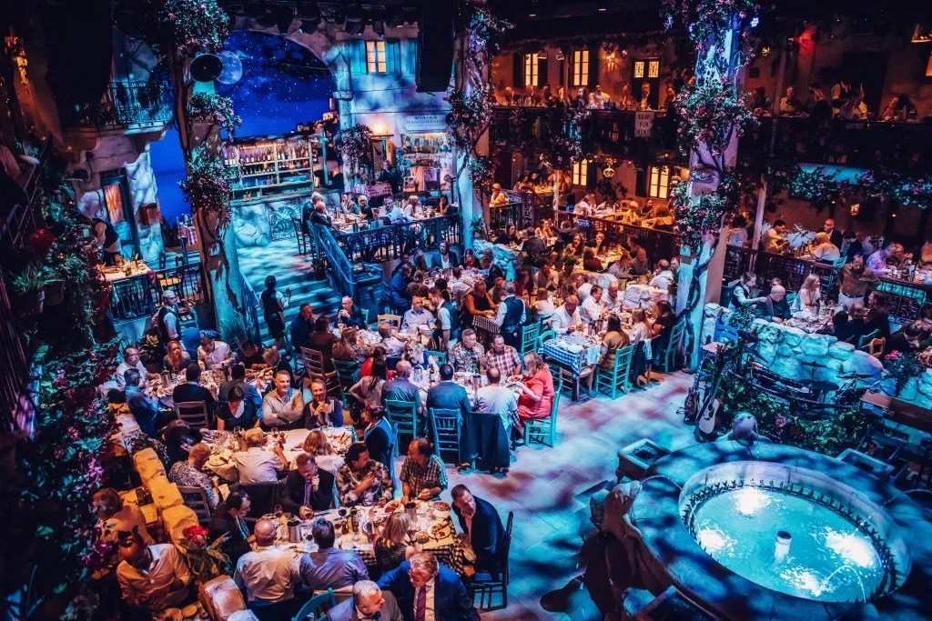 Looking down on the Greek taverna set of Mamma Mia! The Party, with diners at long tables, balconies and a fountain
