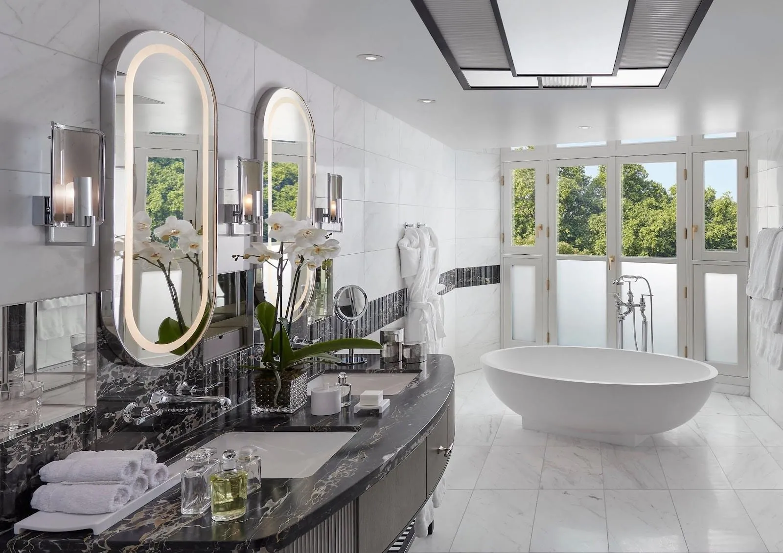 The bathroom of the Two-Bedroom Imperial Suite at Mandarin Oriental Hyde Park: a freestanding oval bath in front of French windows onto trees, twin basins on a black marble counter and illuminated oval mirrors