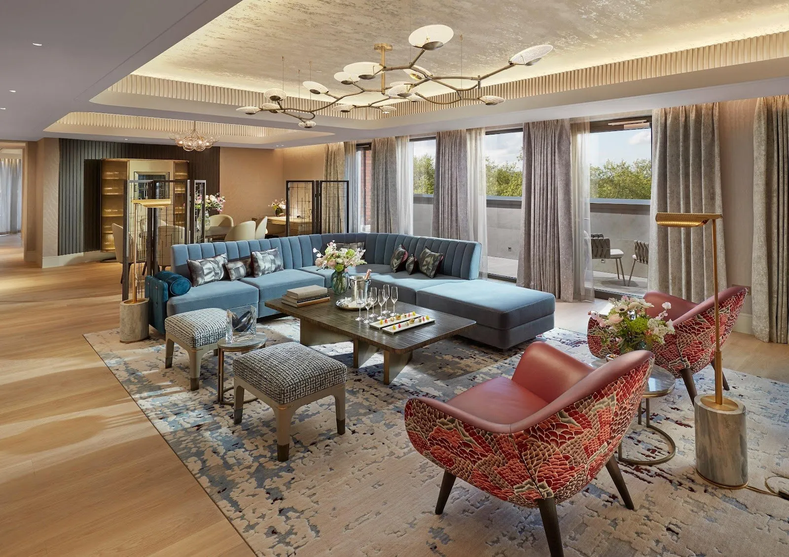A suite lounge at Mandarin Oriental Hyde Park with a blue sectional sofa and a patterned rug, glass doors open to a private terrace with trees beyond