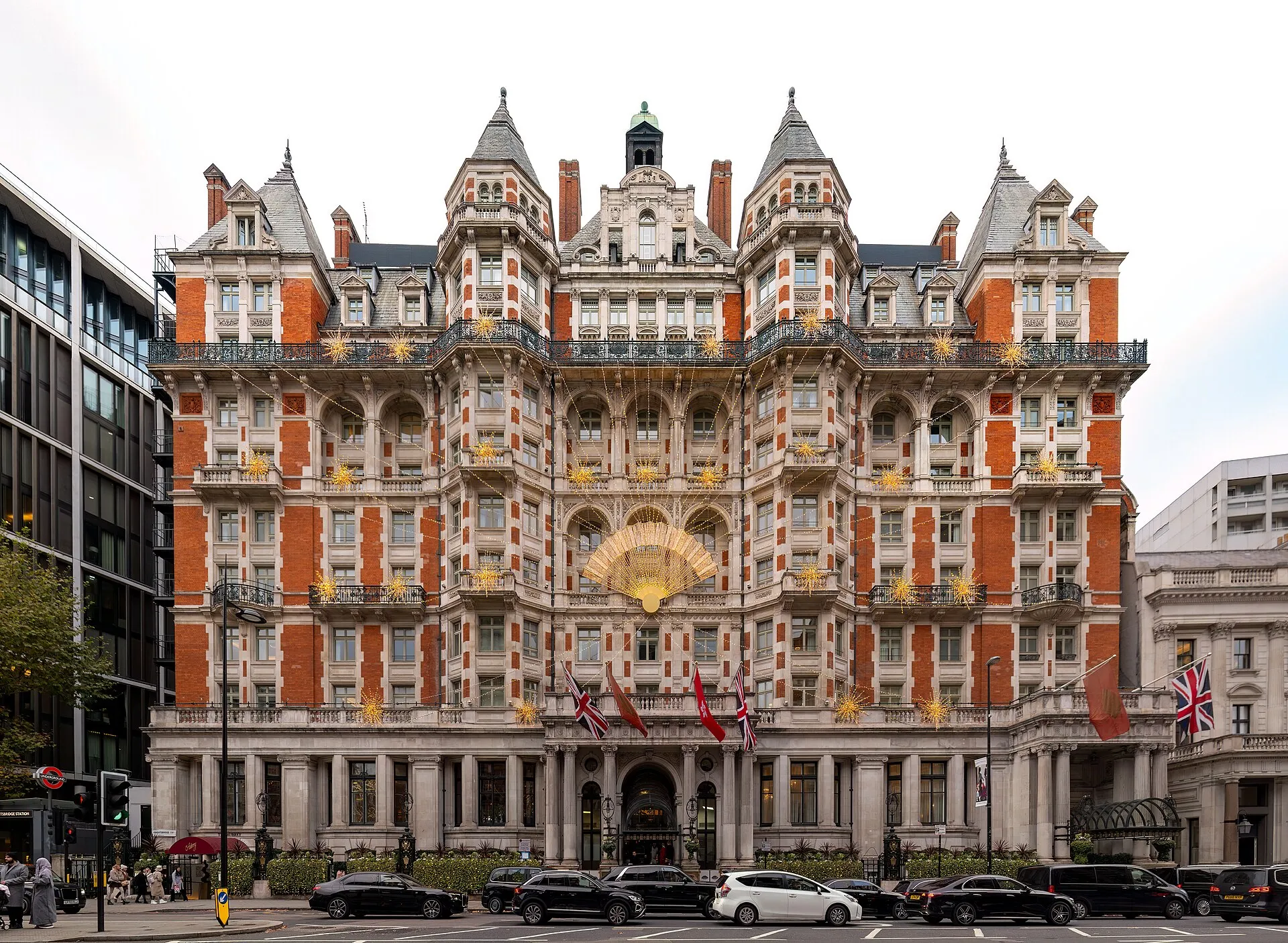 The red-brick and stone front of Mandarin Oriental Hyde Park on Knightsbridge, with turreted roofs, iron balconies, a large gold fan above the entrance and flags over the door, and the Underground roundel of Knightsbridge station at the far left