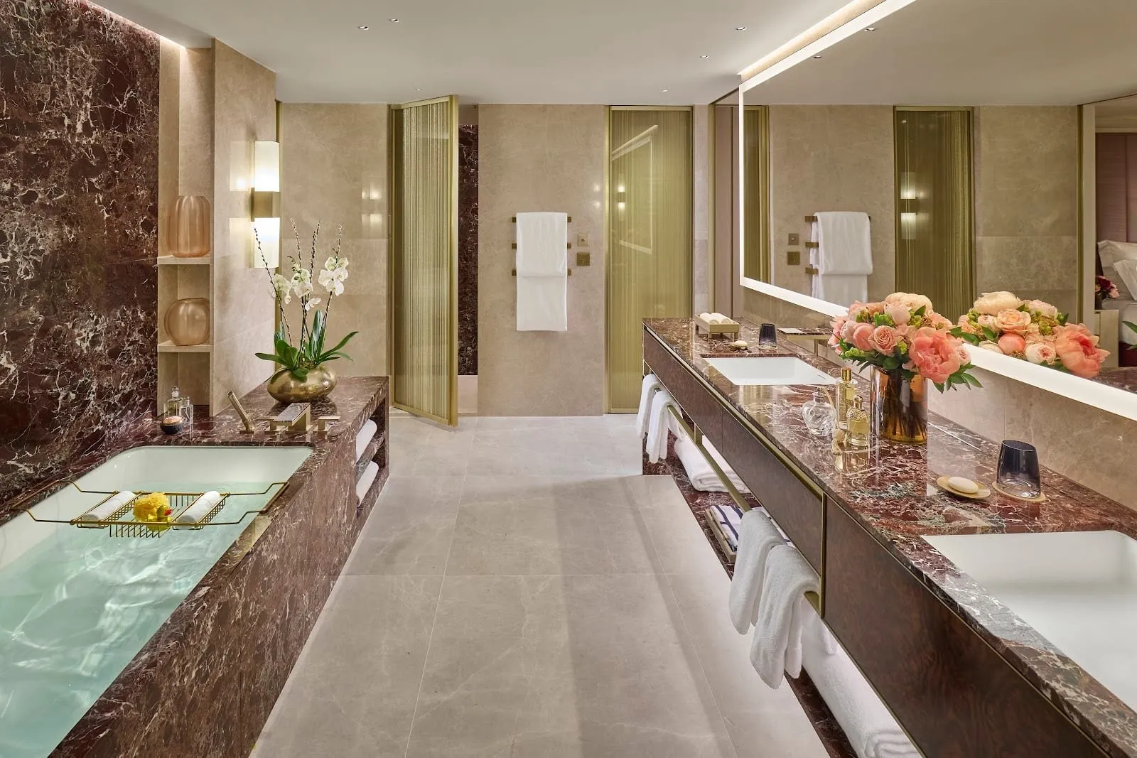 A Mandarin Oriental Mayfair bathroom in red-brown and grey marble, with a bath set in marble, a long twin-basin vanity under a lit mirror and gold mesh doors