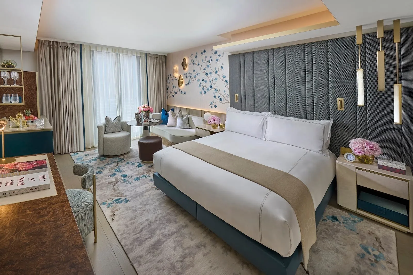 A Mandarin Oriental Mayfair room with a hand-painted blue magnolia panel on the wall, a padded grey headboard with brass lights, a teal bed base, a curved sofa and armchair by the window and a burr-walnut bar