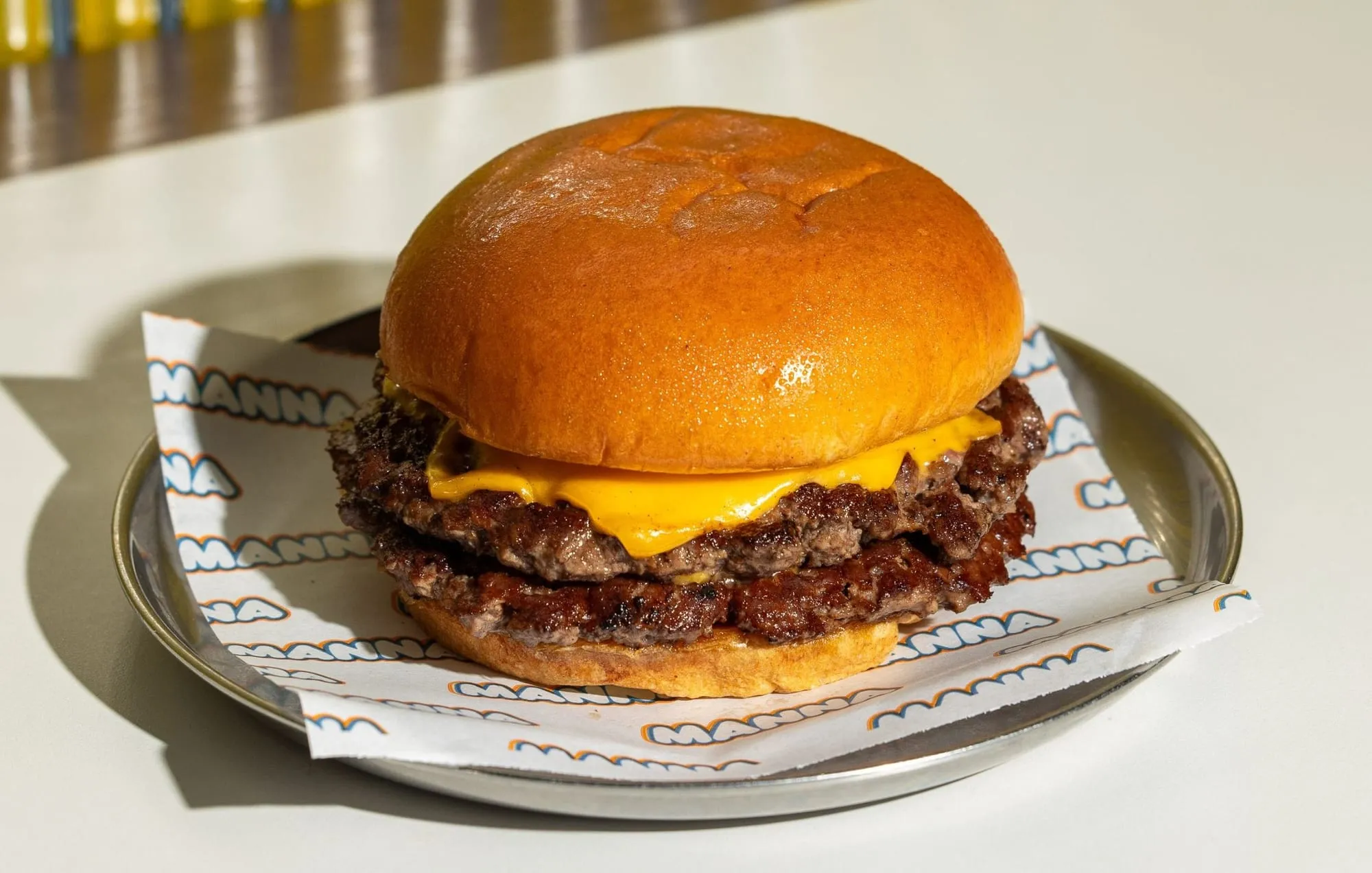 A double cheeseburger on a metal plate, American cheese melting over two thin seared patties in a glossy bun, sitting on greaseproof paper printed with the Manna name