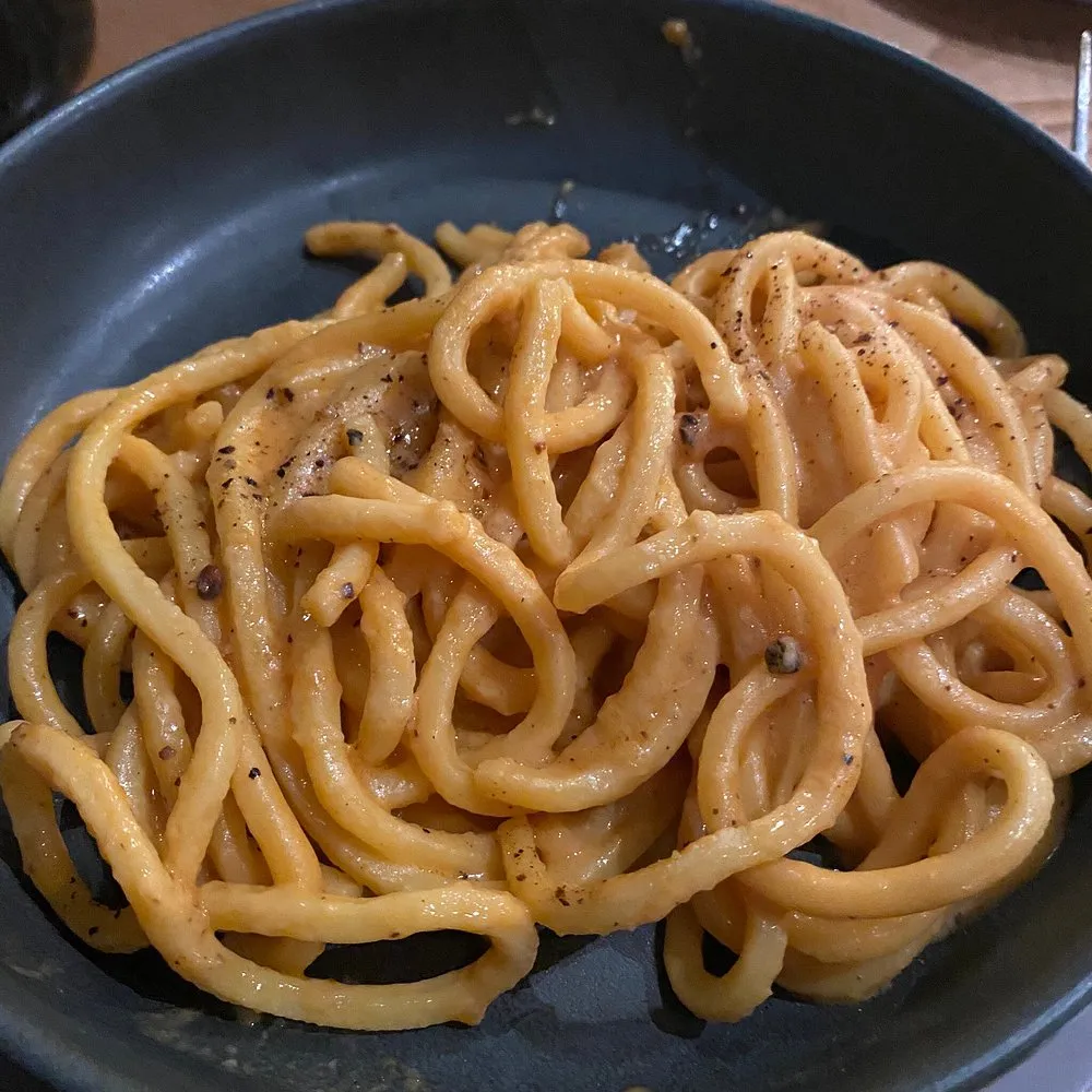 Thick strands of pasta coated in a glossy orange sauce with cracked black pepper, in a dark pan