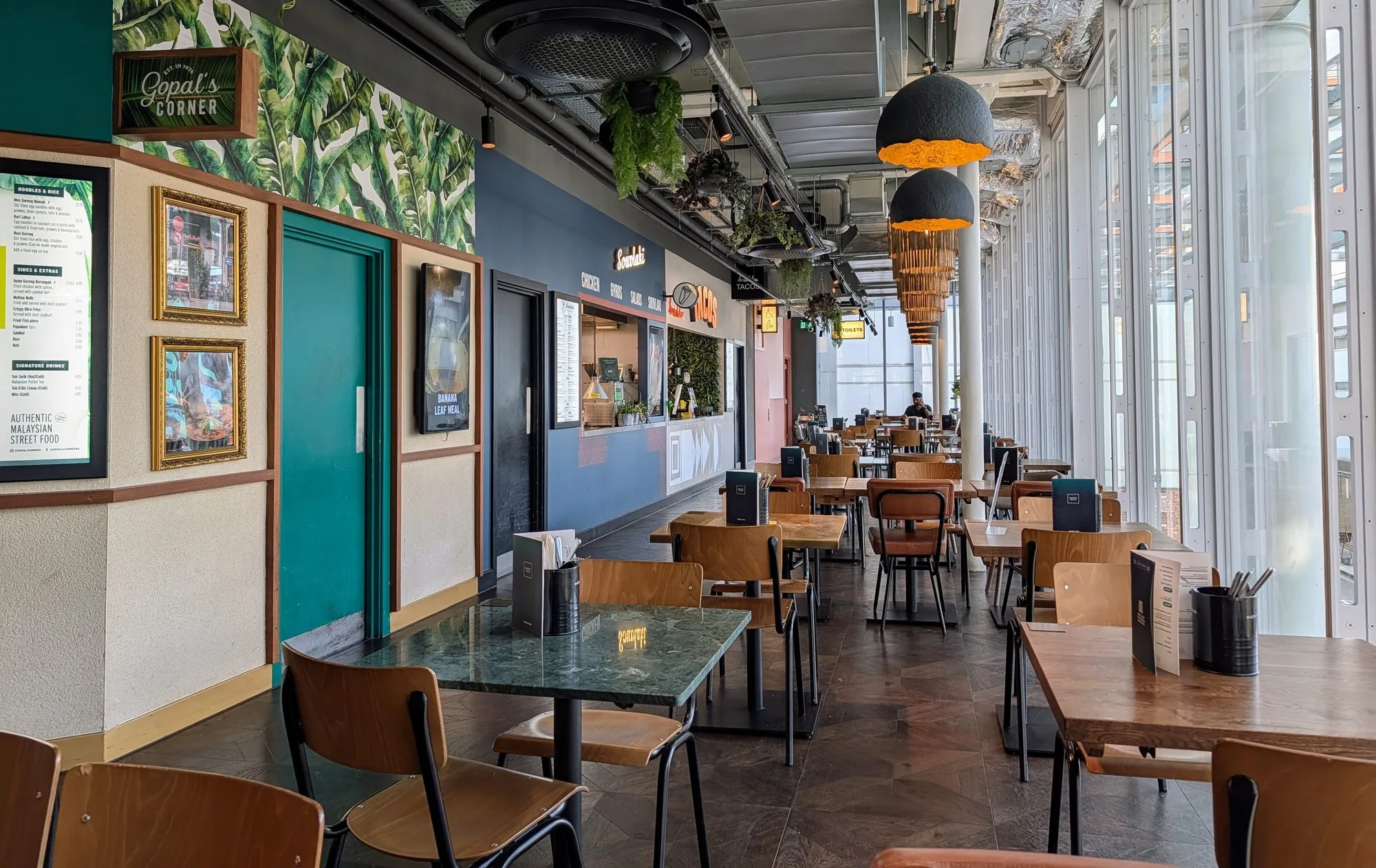 The upper floor of Market Halls Paddington, with the Gopal's Corner Malaysian counter under a banana-leaf mural on the left, a souvlaki and taco counter beyond it, and long communal tables running along a full-height glass wall