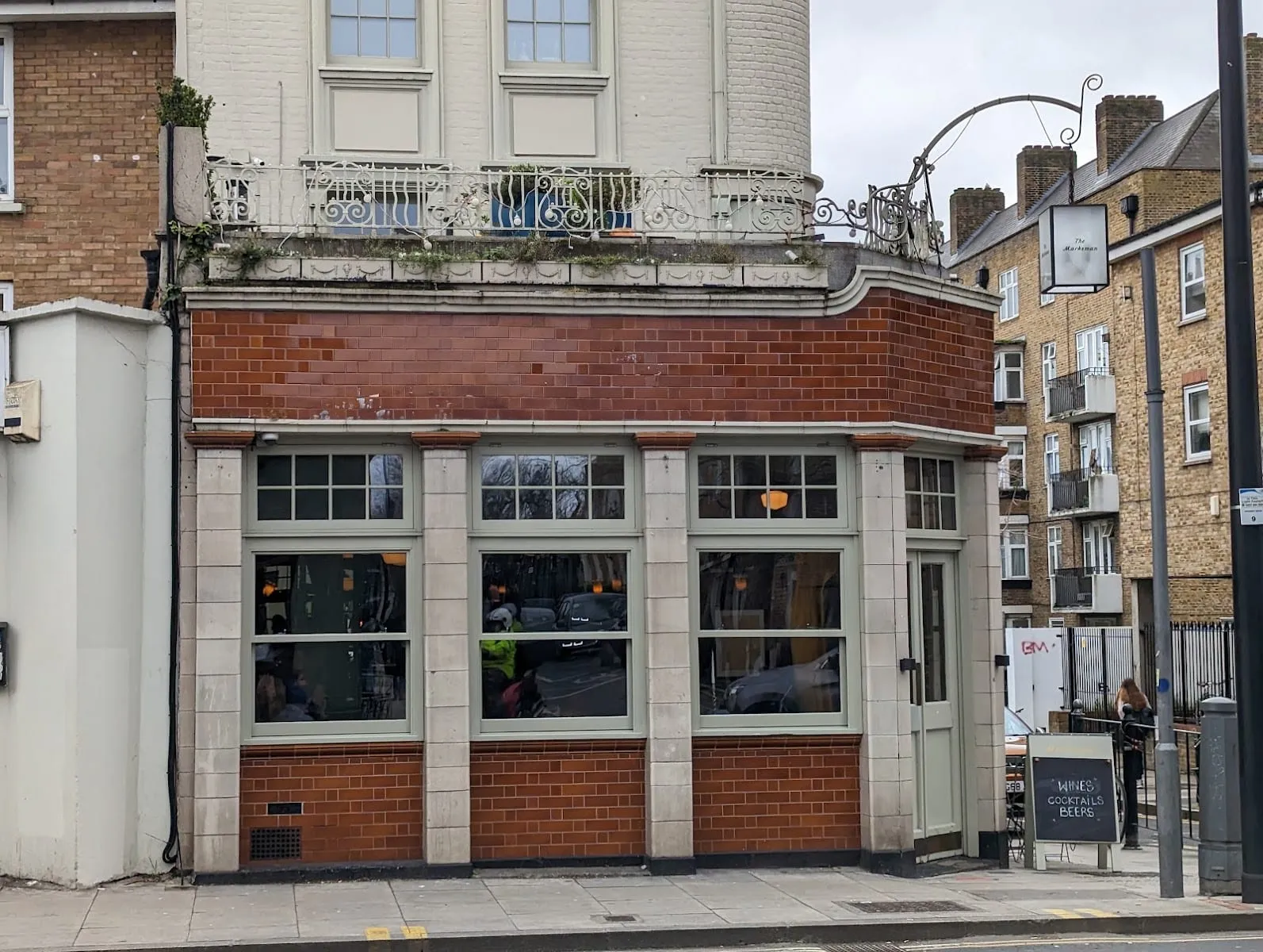 The Marksman on Hackney Road seen from the street: a corner pub in glazed red brick with sage-green windows, a white ornamental balcony above the ground floor, and the pub's hanging sign to the right