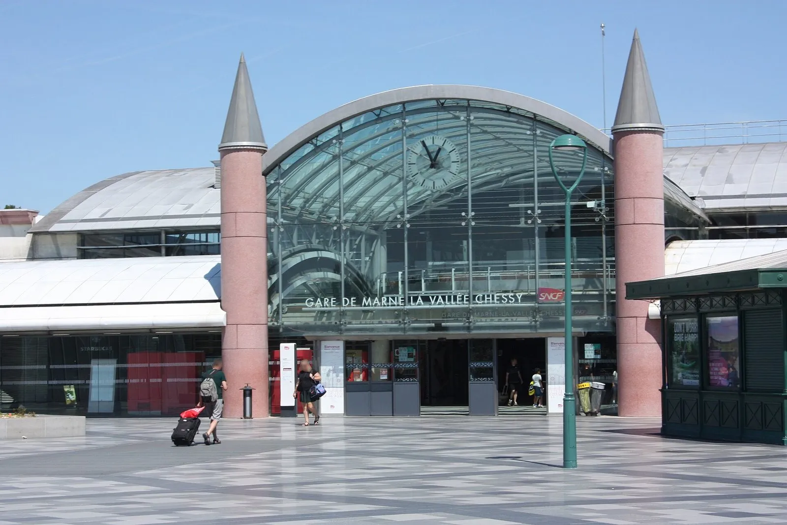The glass-fronted entrance of Marne-la-Vallée–Chessy station, flanked by two pink round towers with grey conical roofs, with a clock above the doors and a paved square in front