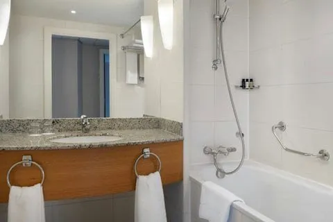 A bathroom at Marriott Executive Apartments Canary Wharf with a bath and shower over it, a grab rail on the tiled wall, and a granite-topped vanity over a wood cabinet