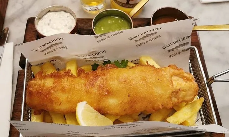 The Mayfair Classic served in a metal frying basket lined with branded paper, with chips, lemon, and pots of tartare sauce, mushy peas and curry sauce alongside