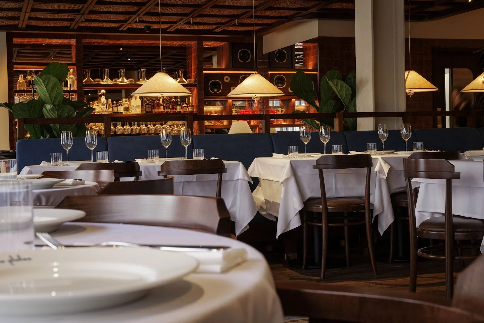 Maza's dining room, with white tablecloths, dark wood chairs, a navy banquette and pleated lampshades in front of a lit back bar